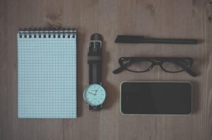 A notepad, watch, pen, pair of glasses, and phone neatly laid out on a wooden surface