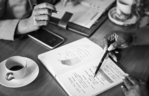 Image of table and hands. Both people are holding a pen. The one on the right is making notes