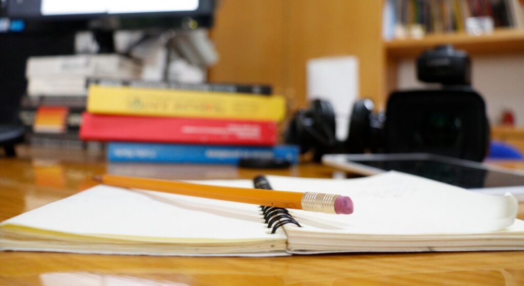An open notebook with a pencil resting on top. On a desk with various office items and books in the background.
Photo credit: Ehécatl Cabrera, CC BY-SA 4.0 <https://creativecommons.org/licenses/by-sa/4.0>, via Wikimedia Commons